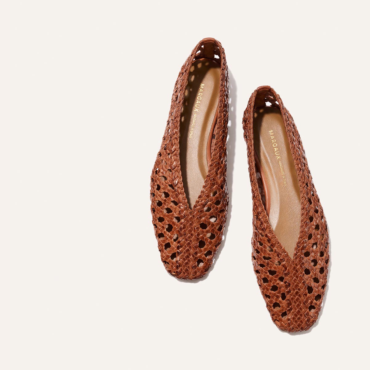 The Woven Paz by Margaux: a pair of saddle brown woven leather flats with pointed toes, handwoven cutout design, and tan insoles featuring gold-printed branding, displayed on a light background.