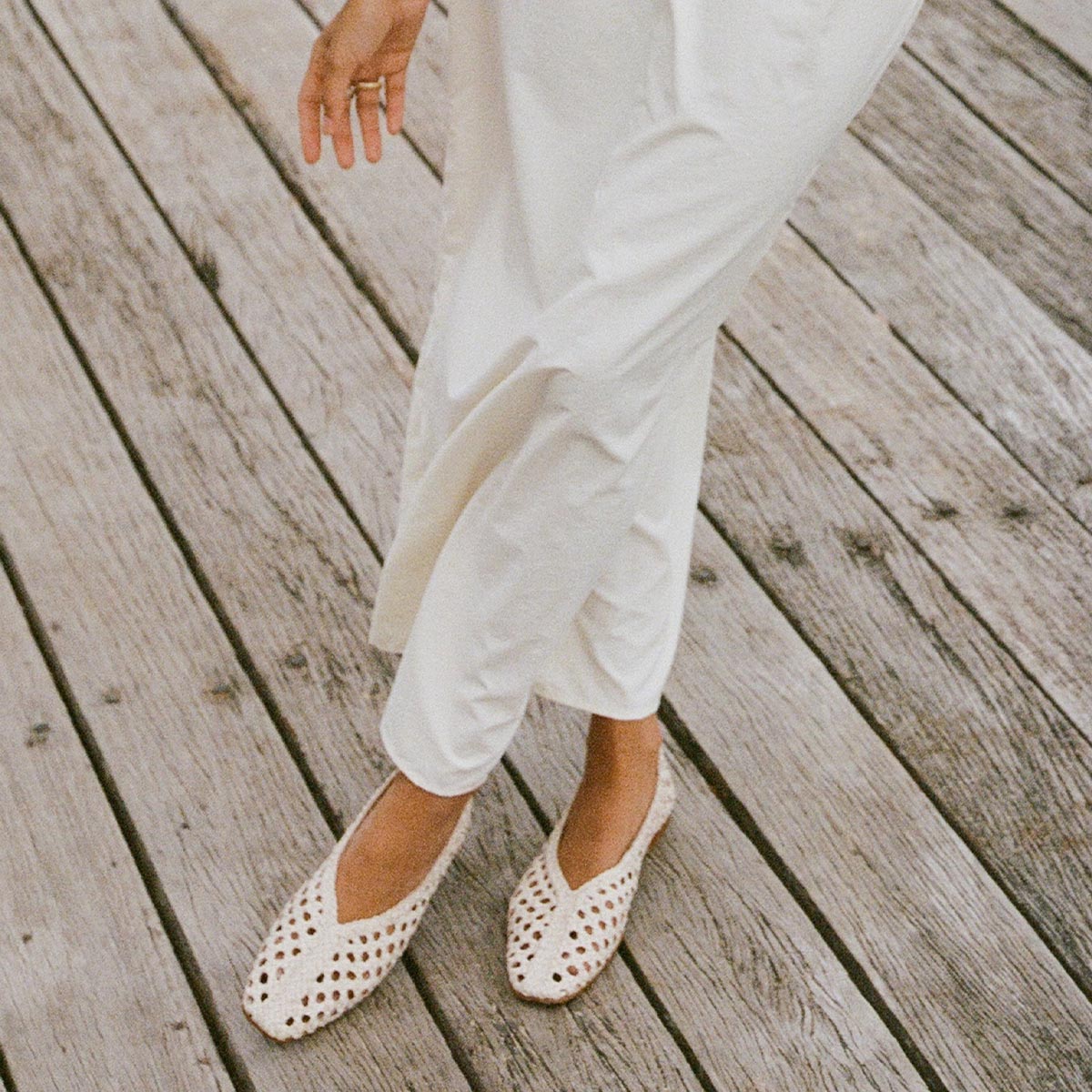 A person wearing high-rise white pants and Margaux’s The Woven Paz SS25 - Ecru Leather shoes stands on a weathered wooden deck, their lower half visible with one hand slightly extended in a relaxed gesture.