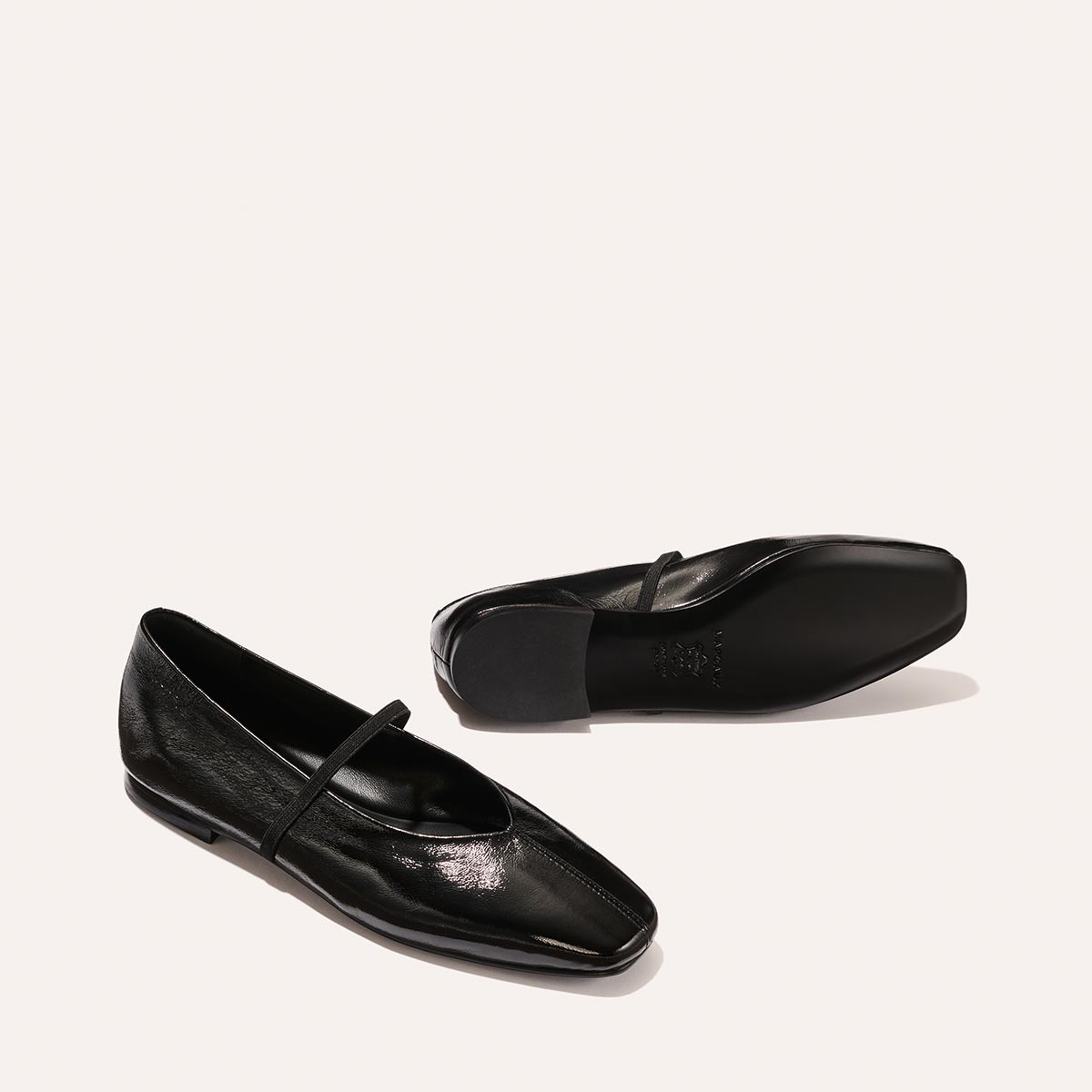 The Phoebe by margaux-development in Black Crinkle-Gloss Nappa: black leather flats with sacchetto construction, a square toe, and a thin strap, displayed on a light background—one shoe upright, the other on its side.