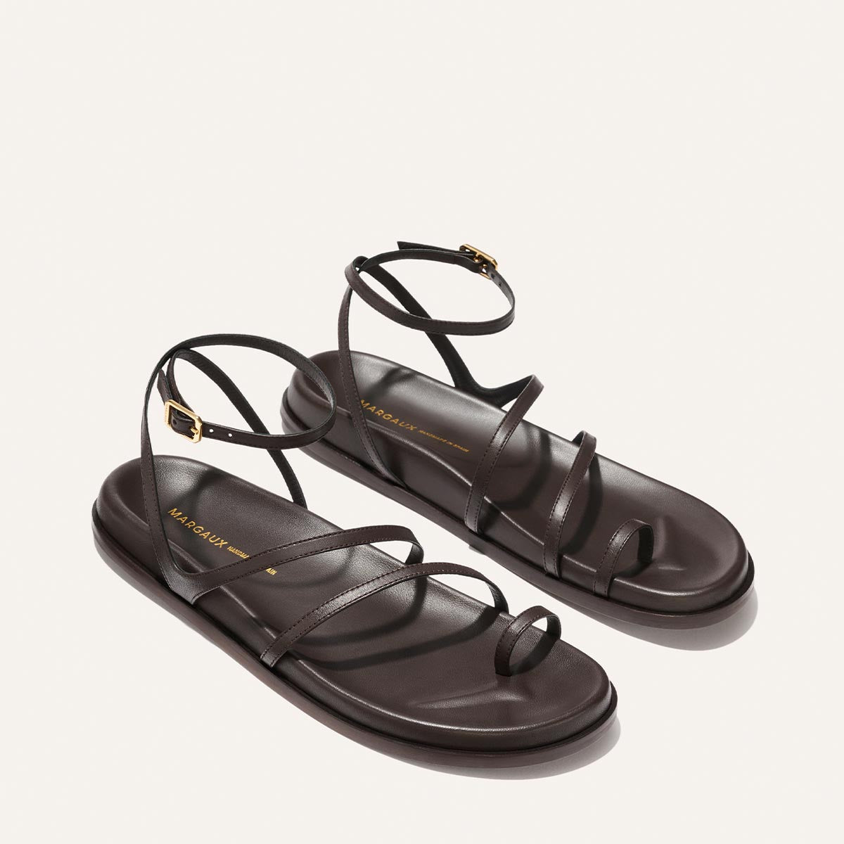 The Vesta Sandal - Espresso Nappa by Margaux: minimalist dark brown leather sandals with thin straps, toe loop, gold-buckled ankle straps, and flat soles, shown on a light background.