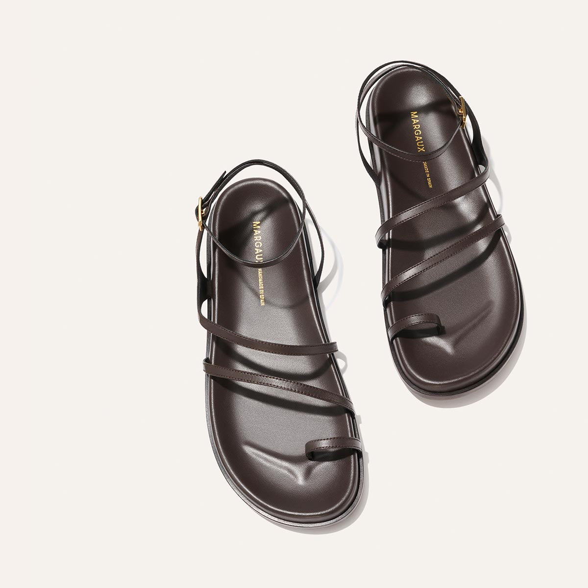 The Margaux Vesta Sandal in Espresso Nappa features thin leather straps, gold buckle details, a minimalist toe loop, and a slightly raised sole, all shown on a light background for a modern look.