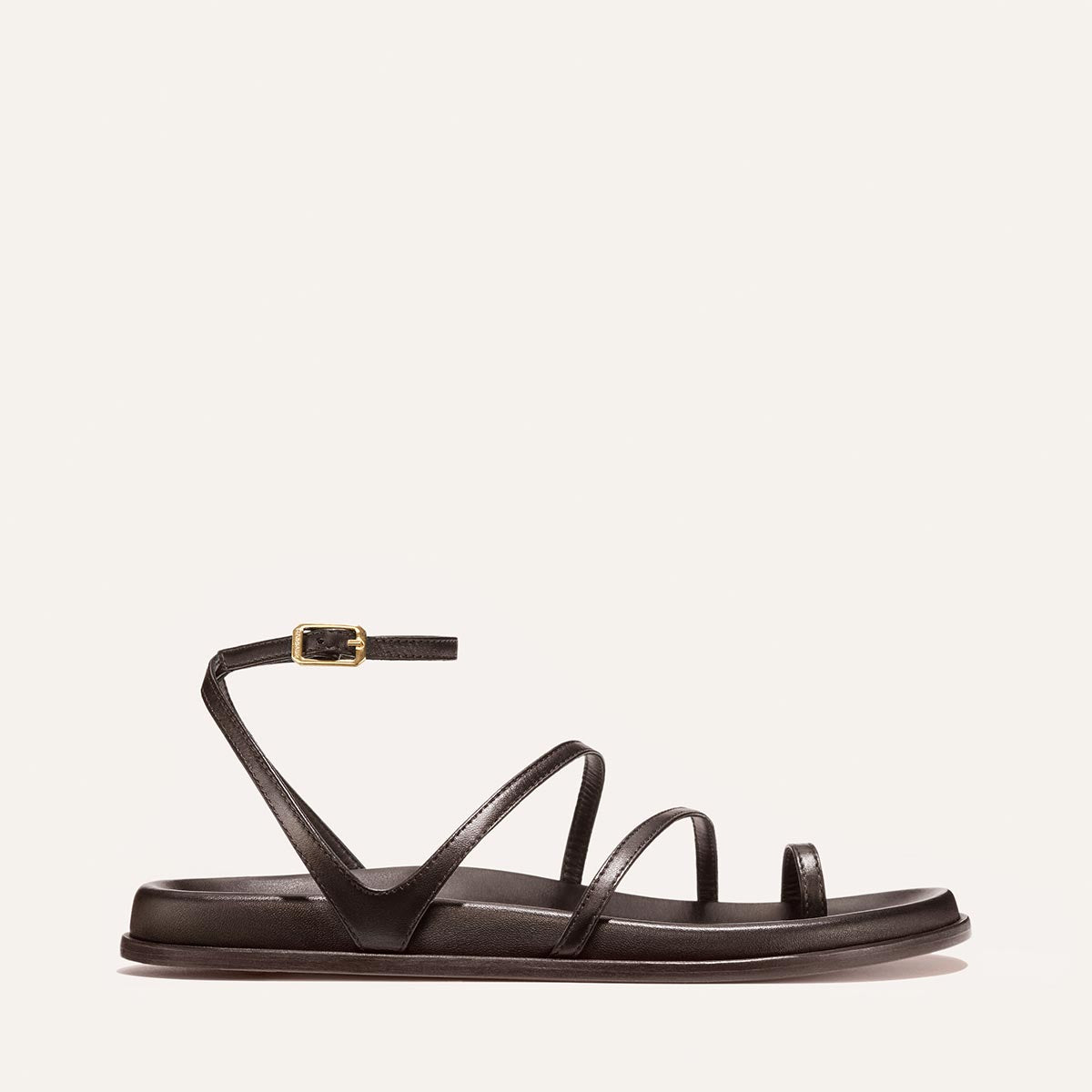 The Vesta Sandal - Espresso Nappa by Margaux features a minimalist flat sole, three thin straps, and an ankle strap with a gold buckle, shown against a plain light background.