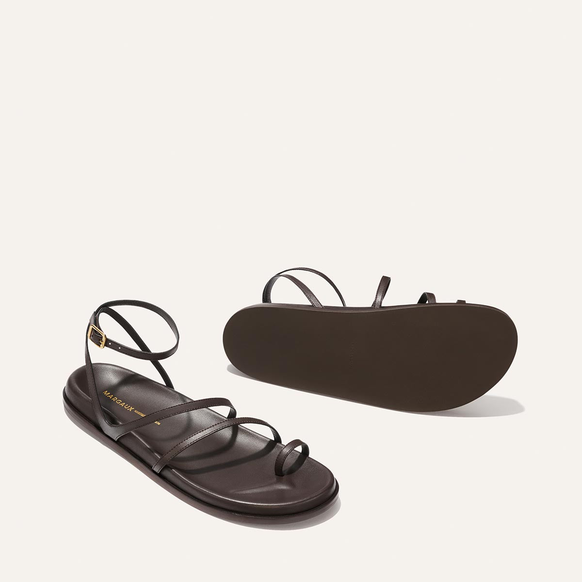 The Vesta Sandal - Espresso Nappa by Margaux: a pair of dark brown, strappy flat sandals with ankle straps. One sandal’s side and the other’s sole show off the minimalist design on a plain background.