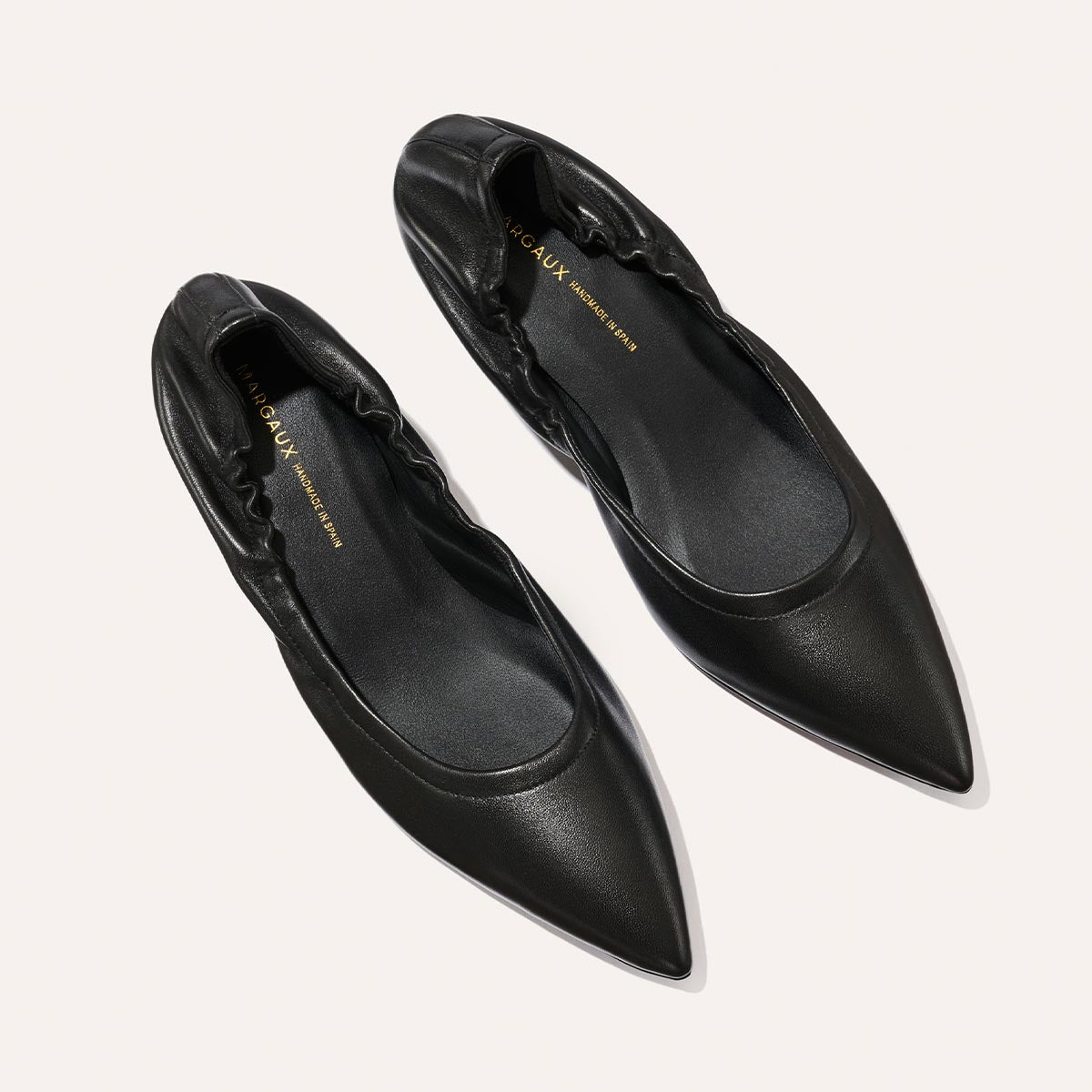 A pair of black Margaux The Roma 40 - Black Nappa pointed-toe flats with gathered detailing and gold-printed insoles, displayed on a light background.