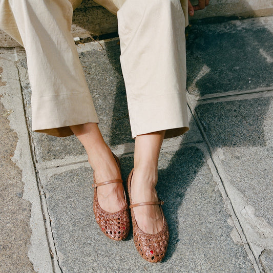 A person in light beige pants wears margaux-developments The Demi Jane - Saddle Woven Leather Mary Jane flats with a cushioned insole, sitting on stone pavement with legs crossed at the ankles.