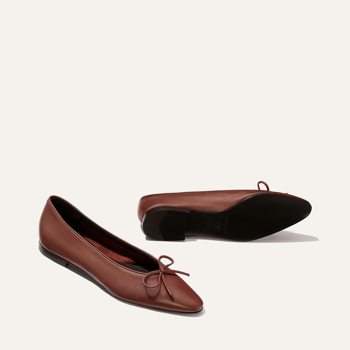 Two “The Salamanca - Brandy Nappa” ballet flats by Margaux, crafted from rich brown nappa leather with pointed toes and delicate bows, are displayed against a plain light background—one standing upright and the other lying on its side to reveal the sleek black sole. These elegant shoes exemplify Margaux’s timeless blend of sophistication and comfort.