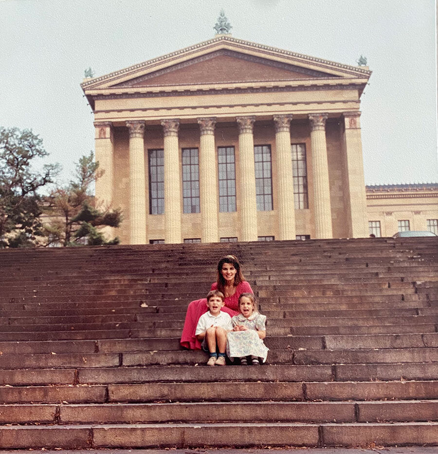 A woman and two children sit on stone steps in front of a large building with tall columns and a triangular pediment, resembling a classical museum or courthouse.