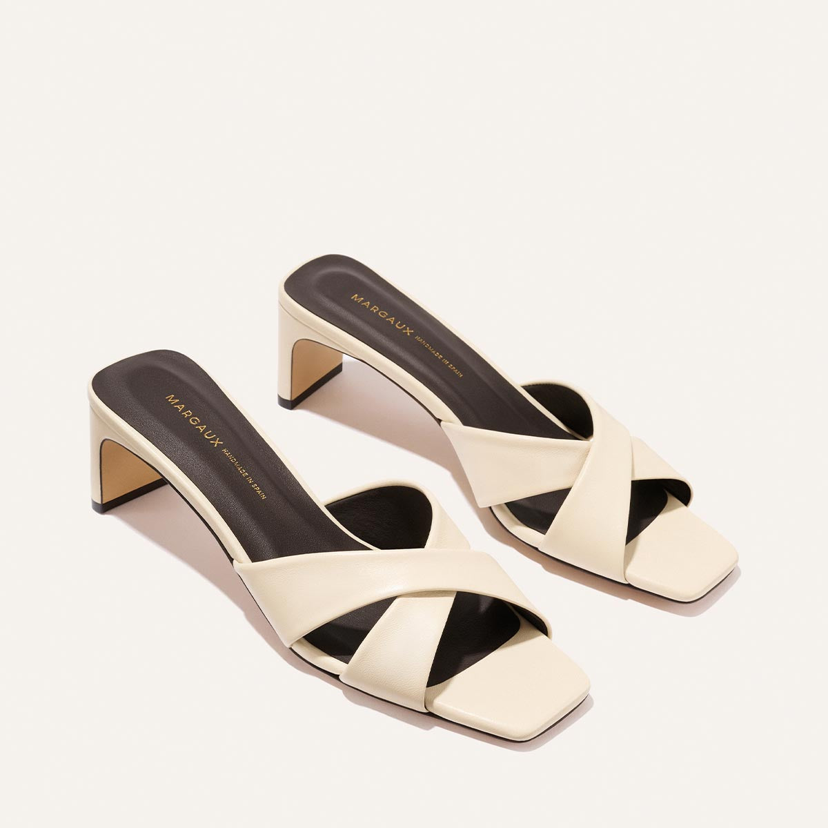The Selene 50 - Ivoire Nappa by Margaux features stylish cream heeled sandals with crisscross straps, open toes, and square heels, shown on a light background.