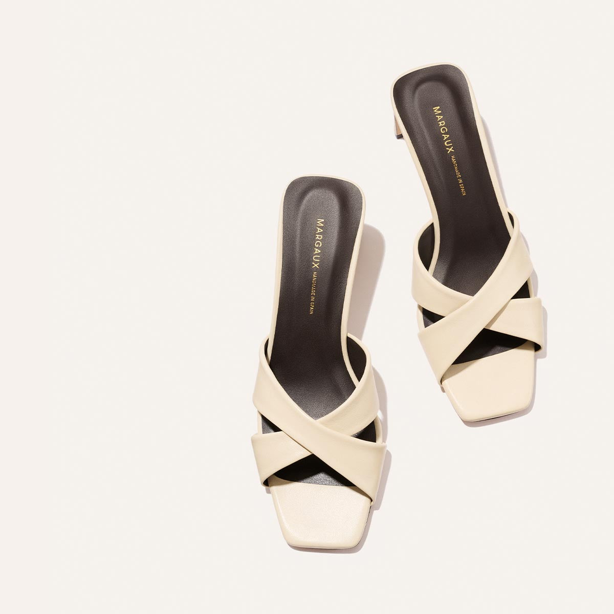 Margaux’s “The Selene 50 - Ivoire Nappa” heeled sandals feature cream crisscross straps and black footbeds, with the brand name in gold on the insoles, arranged diagonally against a light background.