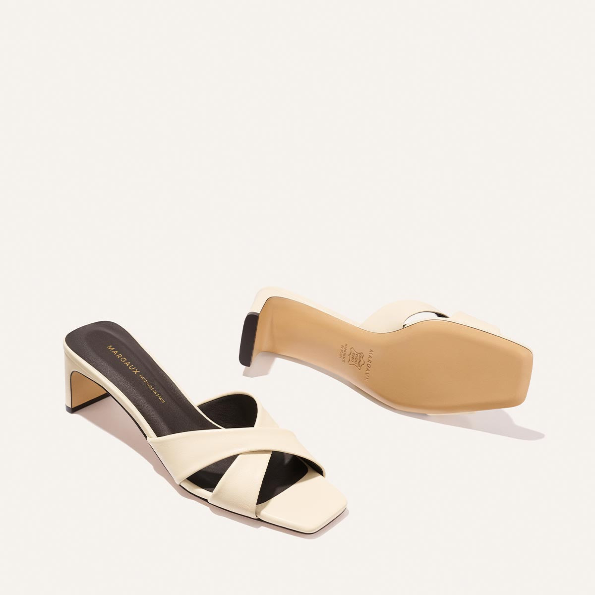 The Selene 50 - Ivoire Nappa by Margaux: a pair of cream open-toe heeled sandals with crisscross straps. One sandal is upright showing the top and side; the other is flipped to reveal the sole.