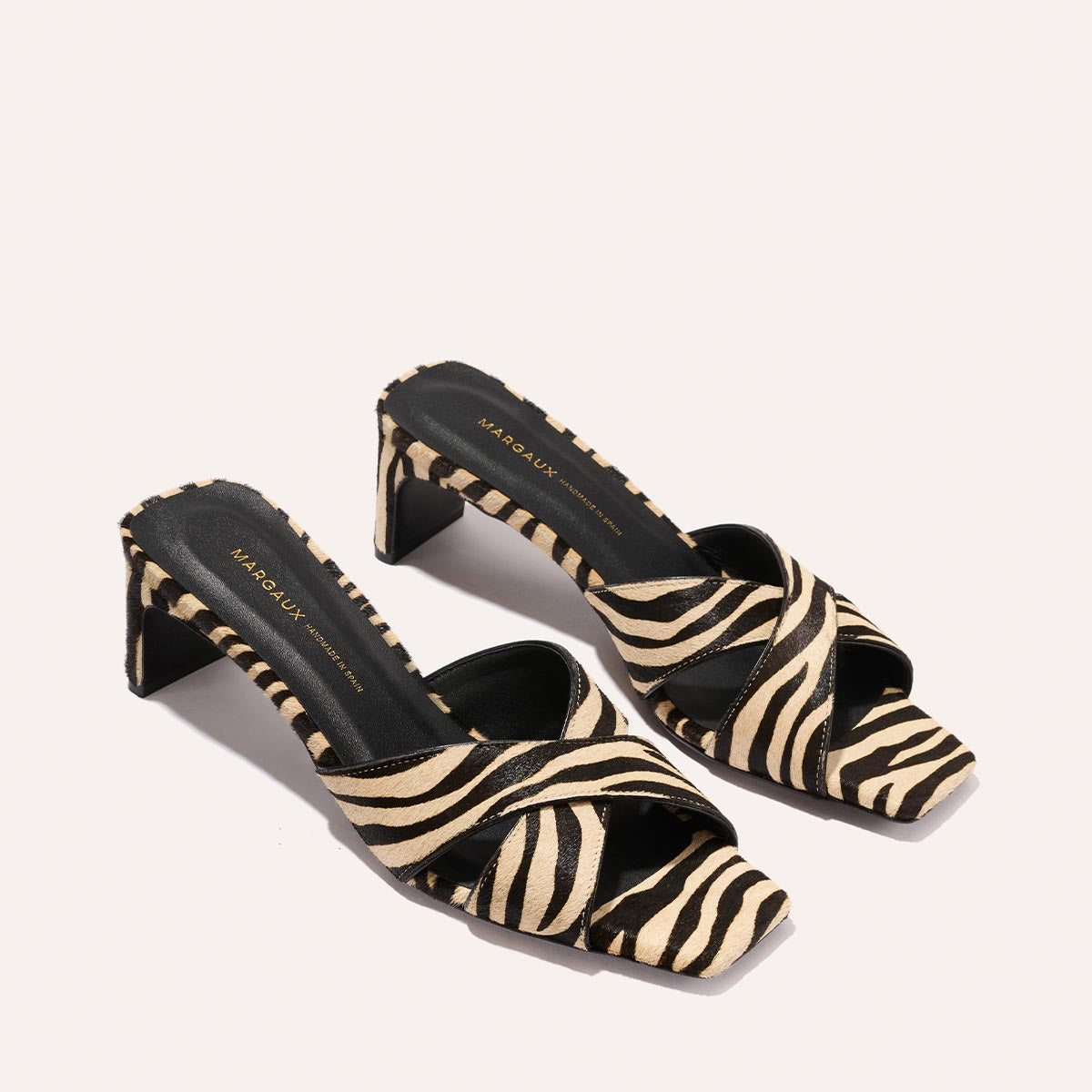 The Selene 50 - Tan Zebra Haircalf by Margaux are open-toe, slip-on block heels featuring black and beige zebra print crisscross straps and a black insole with gold text, set against a light background.