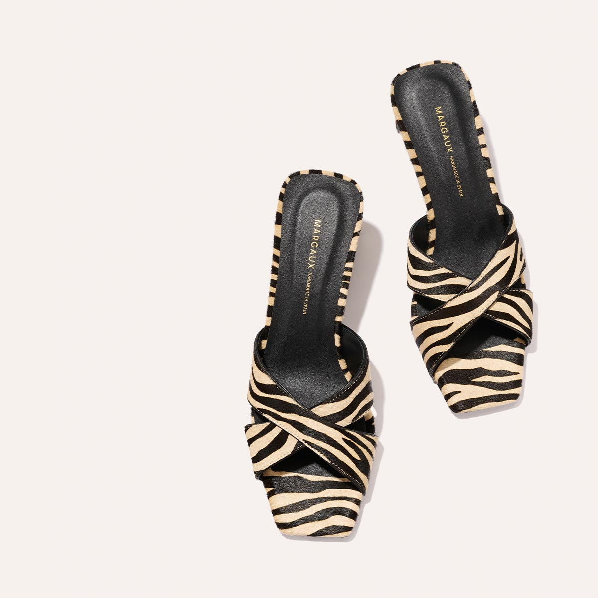 A pair of Margaux The Selene 50 - Tan Zebra Haircalf women's open-toe slip-on shoes with black and beige zebra print, crisscross straps, and black insoles displayed on a light background. The Margaux logo appears on the insoles.