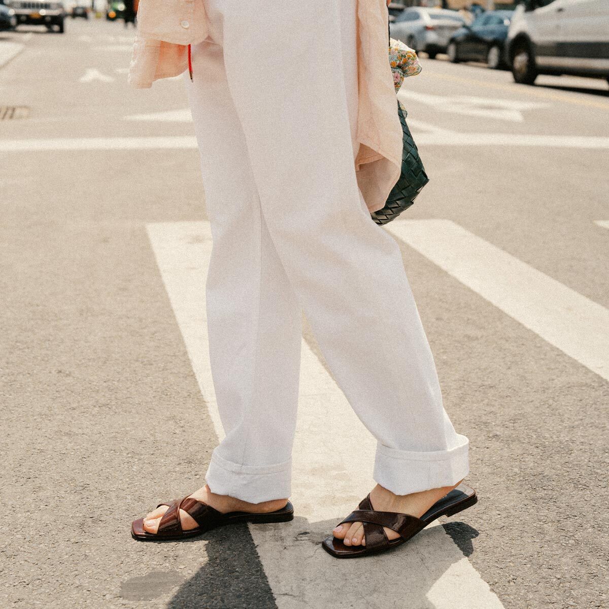 A person stands on a city street in white pants, a pale shirt, and Margaux’s Alex Mill x Margaux: The Selene Flat Sandal in Espresso Crinkle Patent. Their face isn't visible; parked cars and a crosswalk appear in the background.