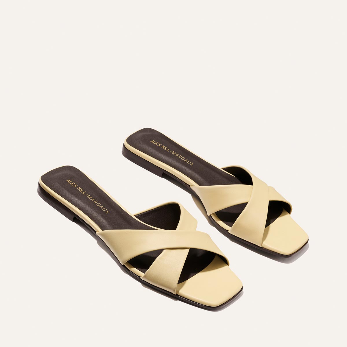 The Margaux Alex Mill x Margaux: The Selene Flat Sandal in Butter Nappa features stylish yellow slip-on design, wide crisscross straps, and flat brown soles, shown on a plain off-white background.