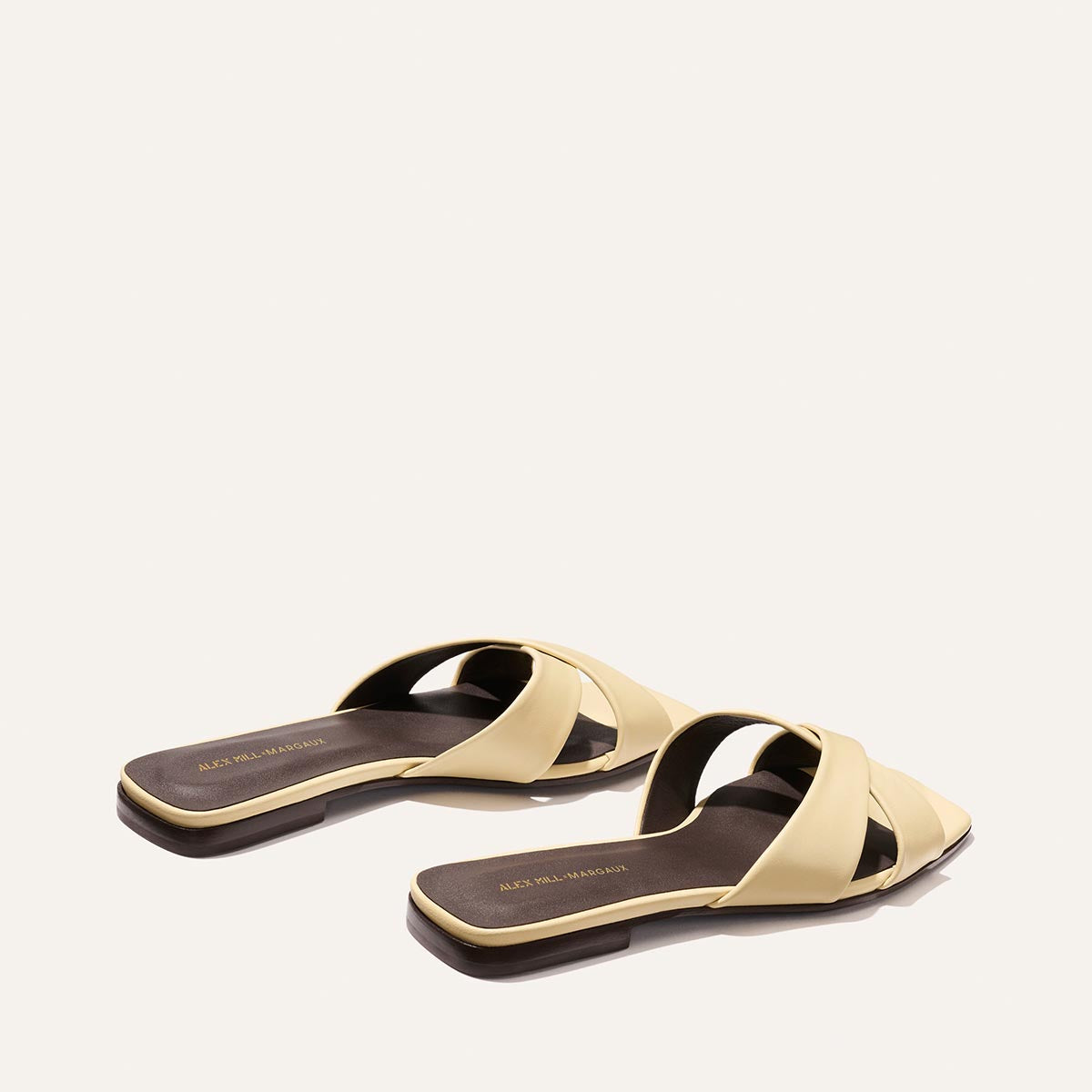 A pair of Alex Mill x Margaux: The Selene Flat Sandal in Butter Nappa by Margaux, featuring pale yellow crisscross straps, open toes, and flat square brown soles, displayed on a plain light background.