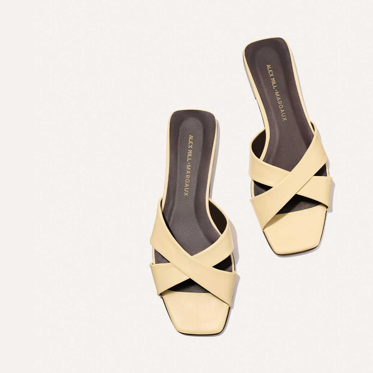 The Margaux Alex Mill x Margaux Selene Flat Sandal in Butter Nappa features pale yellow crisscross leather straps, square toes, and dark insoles with gold branding, all set against a plain background.
