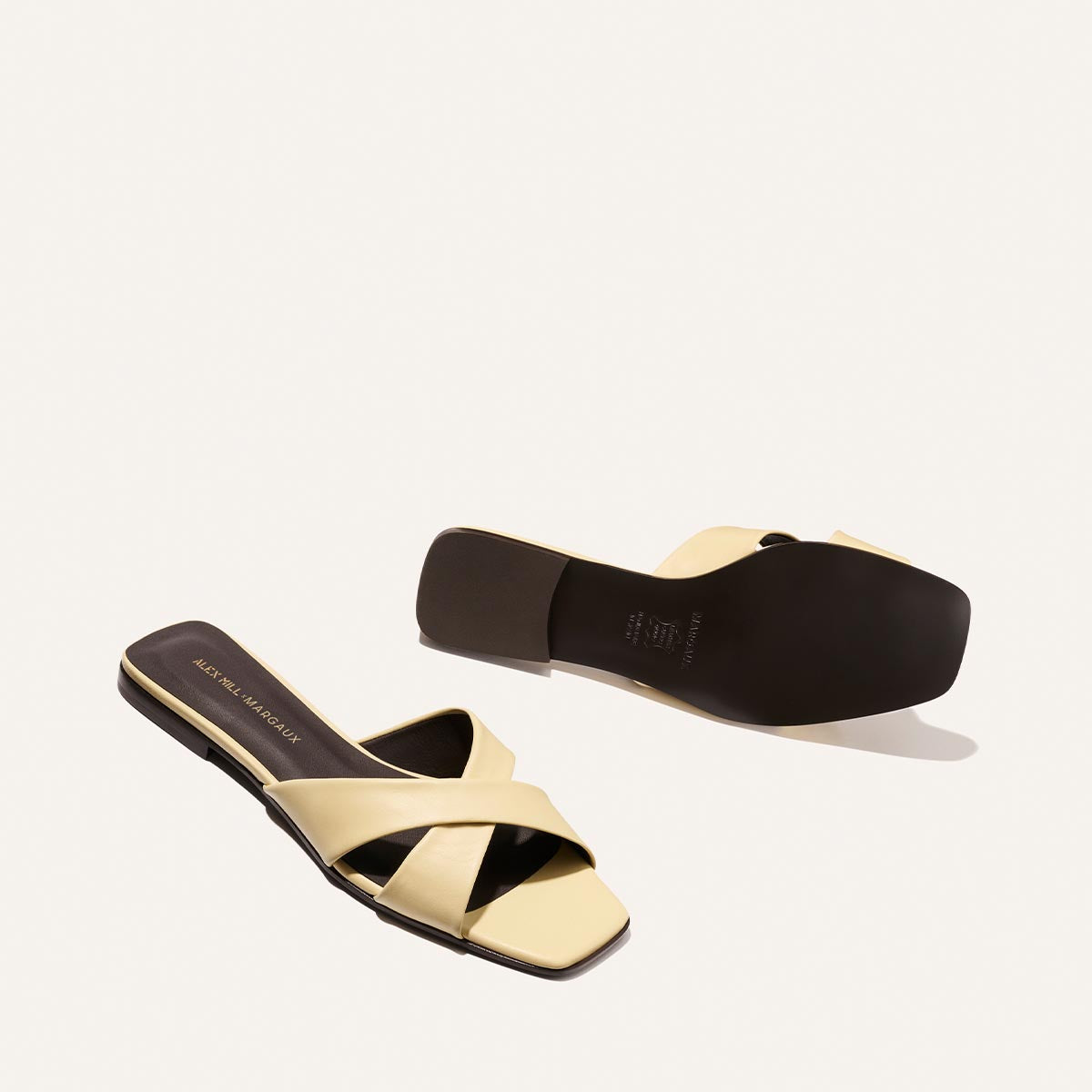 The Margaux Alex Mill x Margaux: The Selene Flat Sandal in Butter Nappa features cream-colored crisscross straps, open toes, and flat black soles—shown from both top and bottom views for a chic summer look.