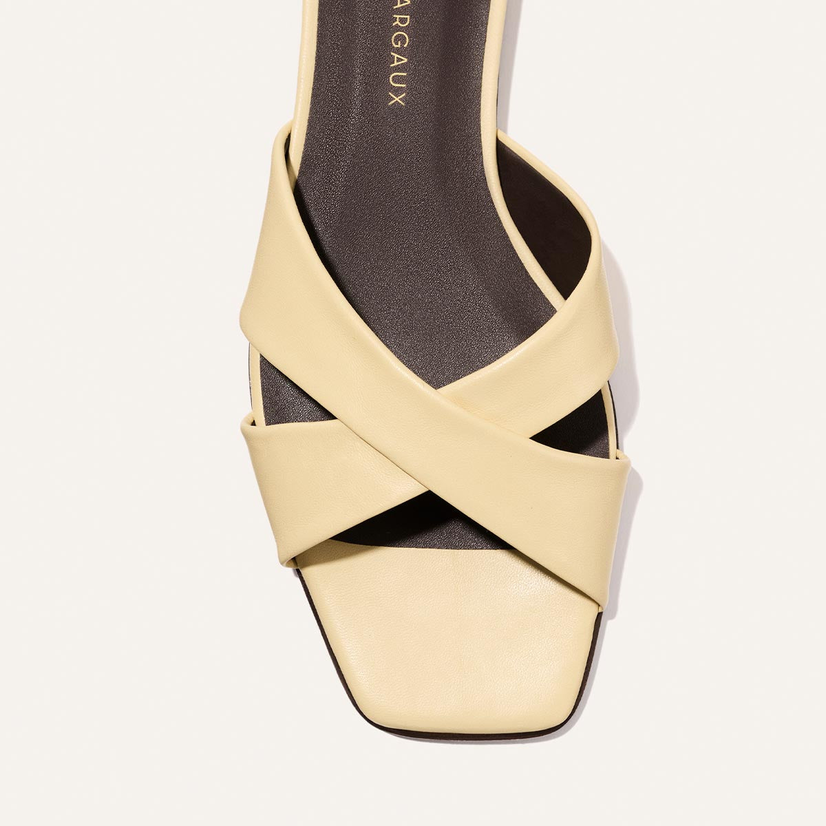 A close-up top view of the Alex Mill x Margaux Selene Flat Sandal in Butter Nappa shows its squared toe, two wide cream-colored crisscross leather straps, and dark brown insole.