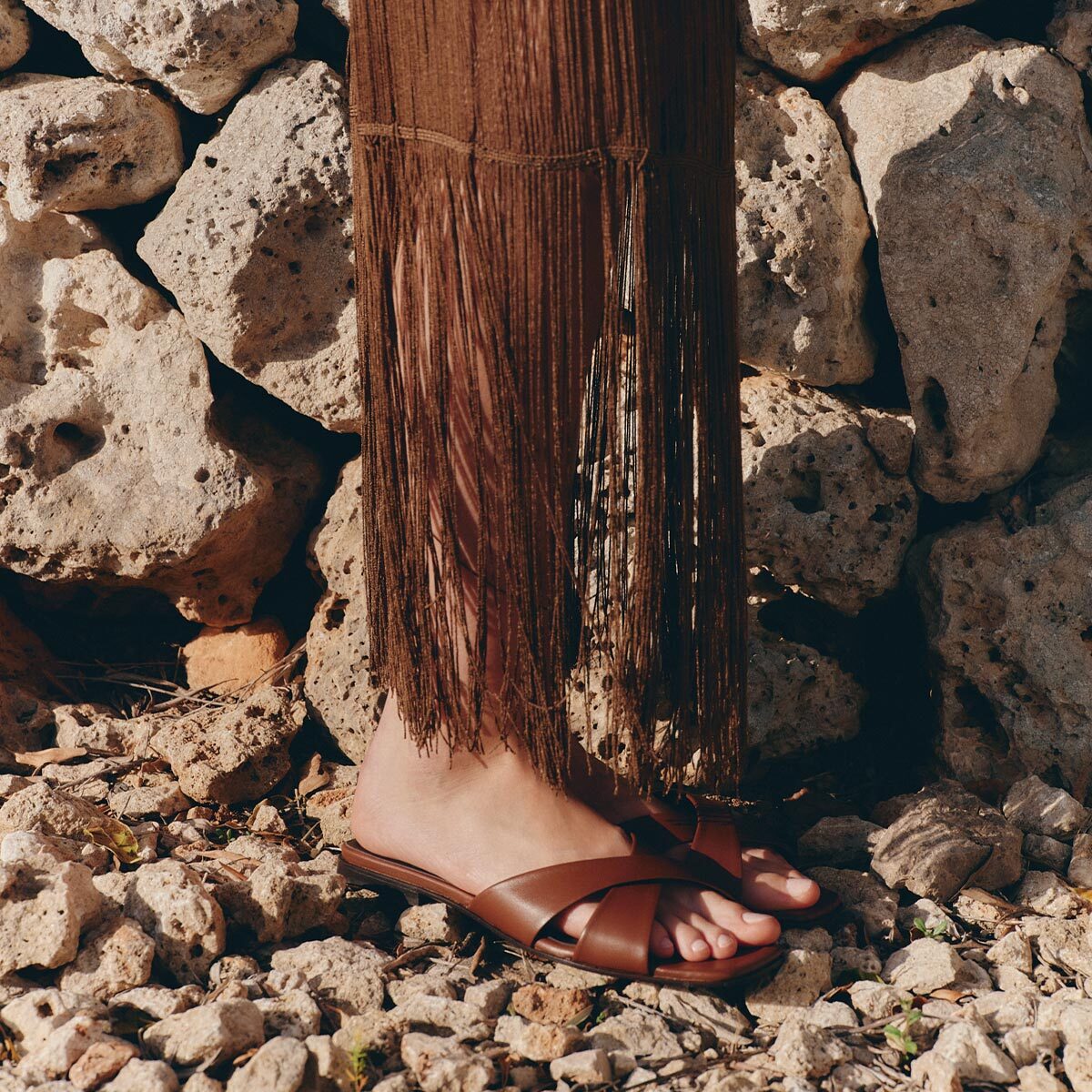 A person stands on rocky ground, wearing Margaux’s The Selene Flat Sandal in Cognac Nappa and a brown fringed skirt, in front of a wall made of large rough stones.