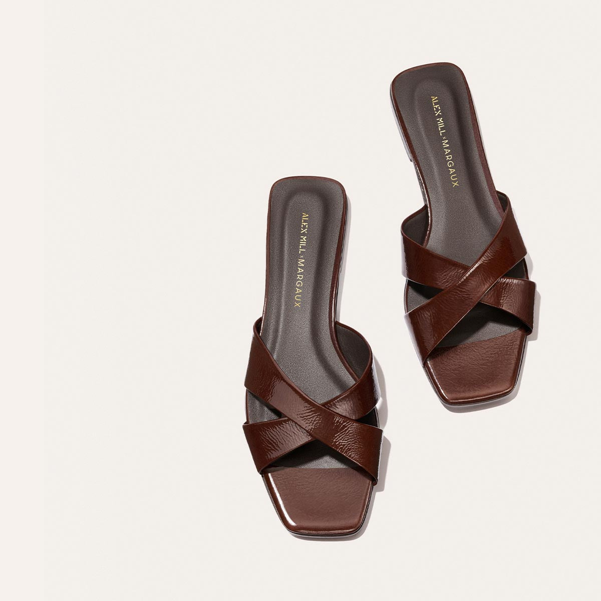 The Margaux Alex Mill x Margaux Selene Flat Sandal in Espresso Crinkle Patent features dark brown, crisscross leather straps, square toes, and a black insole with gold text—ideal for a chic summer look.