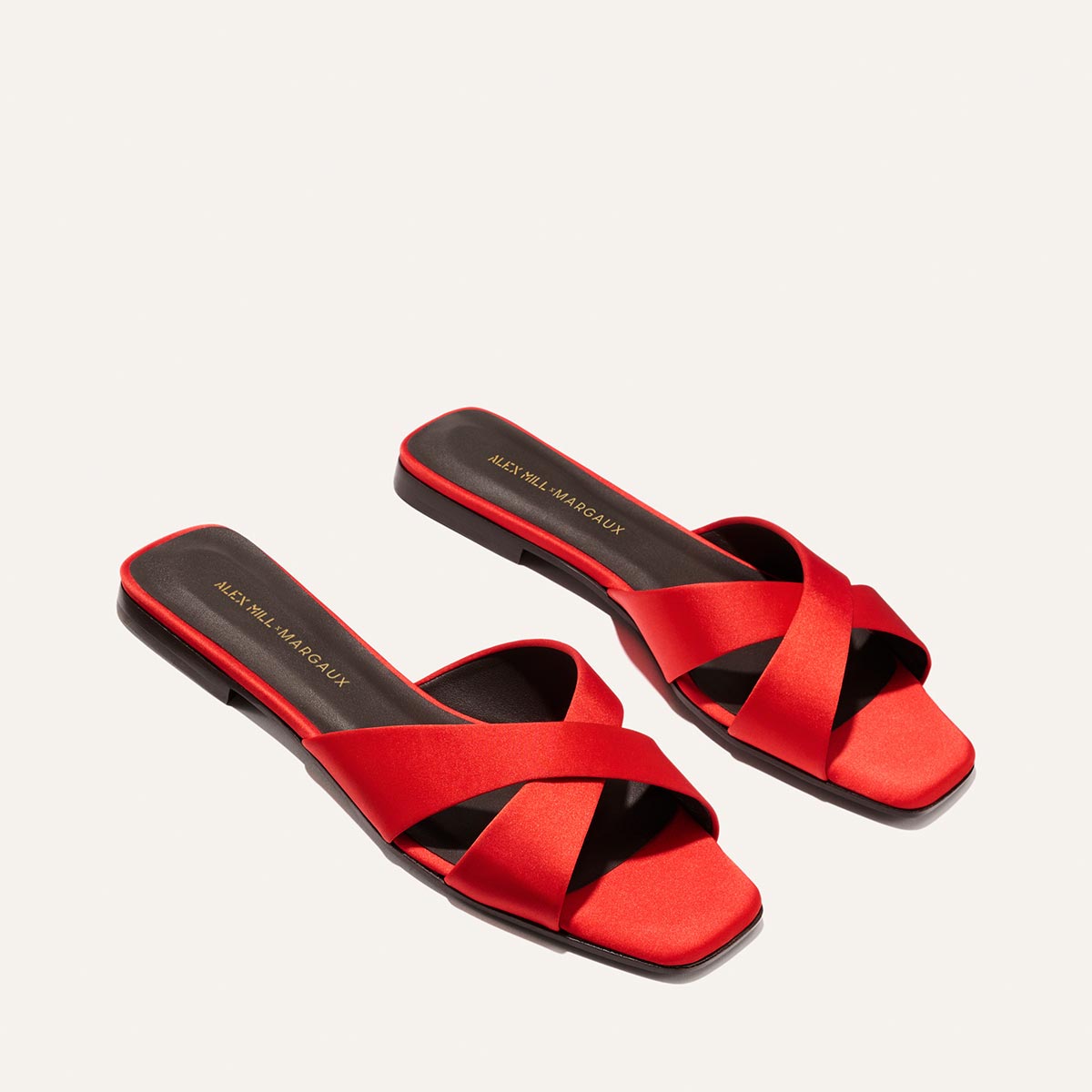 The Alex Mill x Margaux: The Selene Flat Sandal - Poppy Satin features red satin crisscross straps, square toes, and black insoles with gold text, shown on an off-white background—an effortless summer style by Margaux.