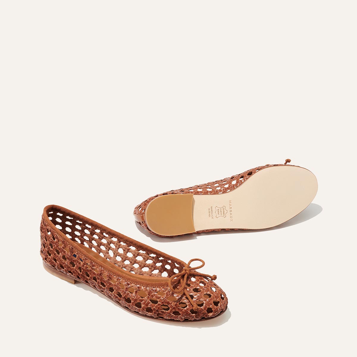 The Demi - Saddle Woven Leather by margaux-development: brown woven leather ballet flats with rounded toes and bows, shown on a light background. One shoe upright, one showing its smooth, light sole. Handmade in Spain.